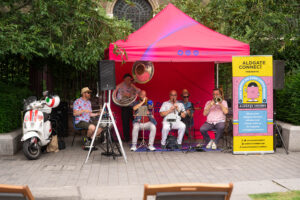 A brass band play music at Aldgate Sounds 2025, to promote the summer listing of Aldgate Sounds 2026